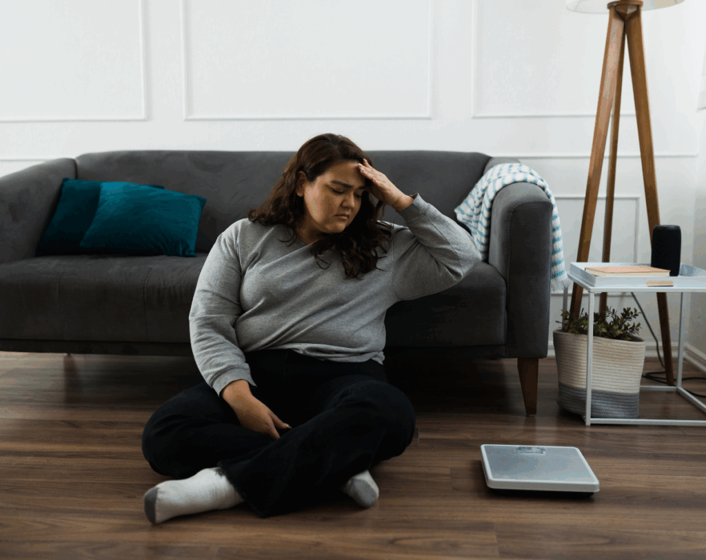 Woman sitting on the floor, looking distressed, in front of a scale, reflecting the emotional struggles of weight loss challenges for women.
