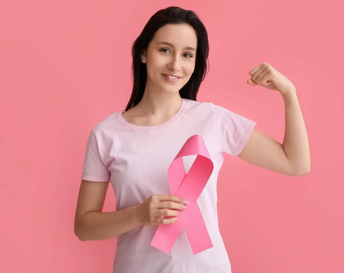 Woman in a pink shirt flexing her arm and holding a pink ribbon, symbolizing breast cancer awareness, against a pink background.