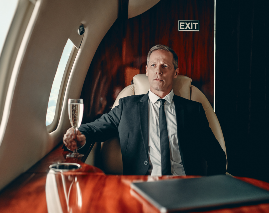 Businessman in a suit holding a champagne glass inside a luxury private jet, with a wooden interior and an exit sign visible.