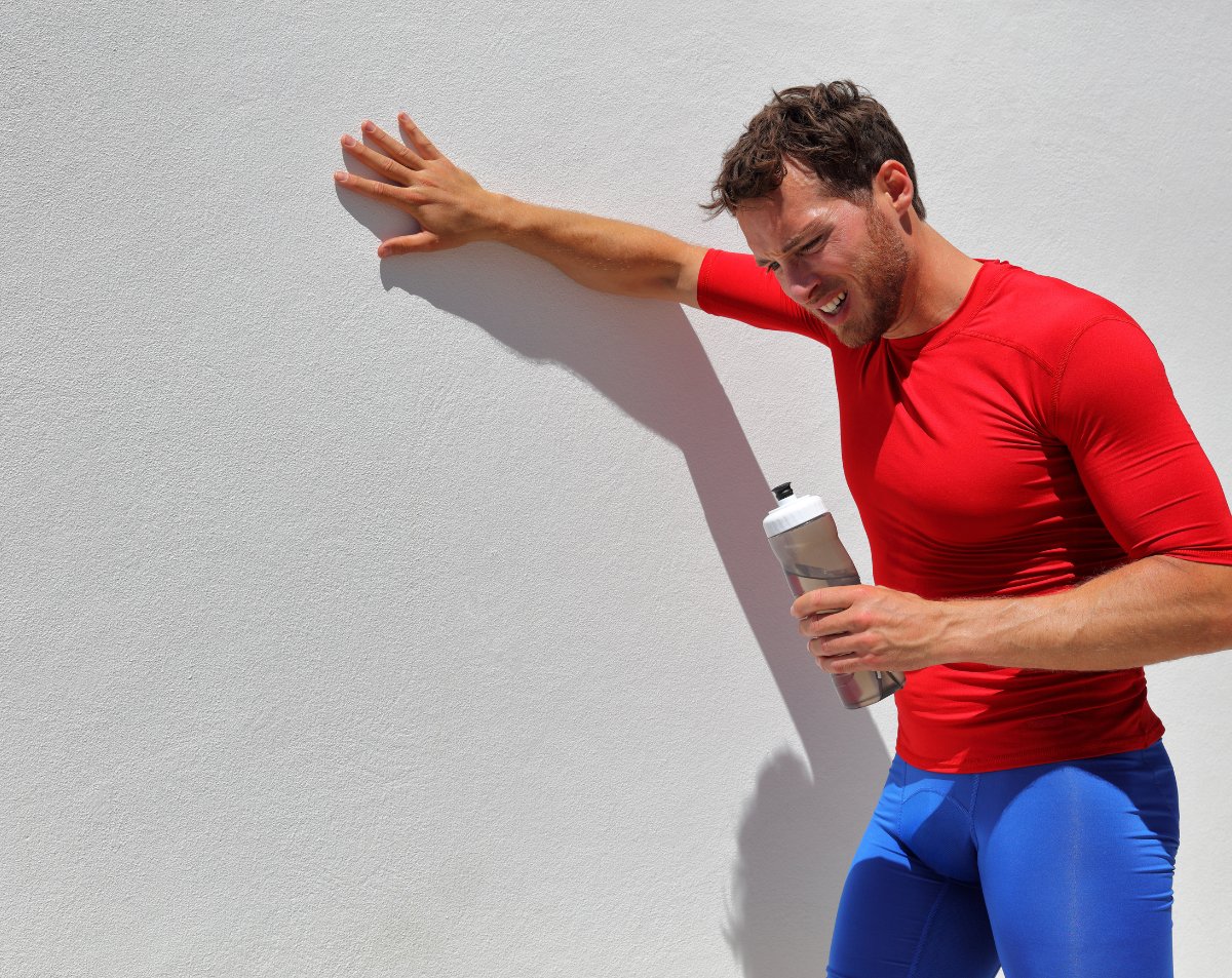 Man in red athletic shirt and blue shorts leaning against a wall, showing signs of dehydration after outdoor activity, holding a water bottle.